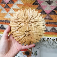 Hand holding a wooden leaf-shaped ornament with a face design against a patterned rug.